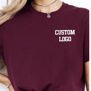 May include: A maroon t-shirt with the text "CUSTOM LOGO" printed in white on the chest. The shirt has a crew neck and short sleeves. The model is wearing a necklace.