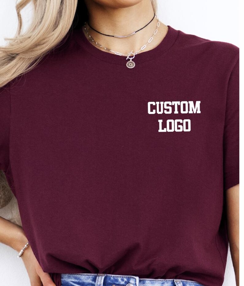 May include: A maroon t-shirt with the text "CUSTOM LOGO" printed in white on the chest. The shirt has a crew neck and short sleeves. The model is wearing a necklace.