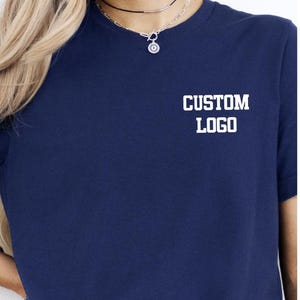 May include: A navy blue t-shirt with the words "CUSTOM LOGO" printed in white block letters. The shirt has short sleeves and a crew neck. A silver necklace with a small pendant is visible.