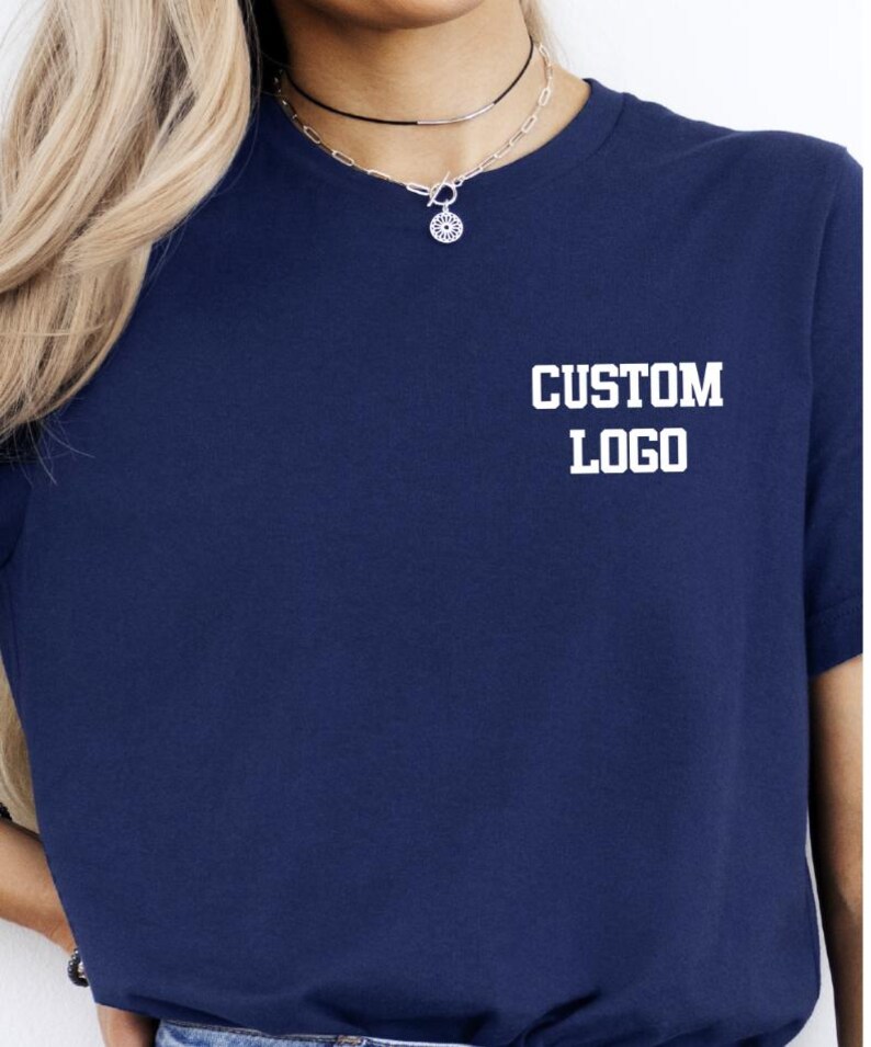 May include: A navy blue t-shirt with the words "CUSTOM LOGO" printed in white block letters. The shirt has short sleeves and a crew neck. A silver necklace with a small pendant is visible.