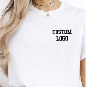 May include: White t-shirt with the text "CUSTOM LOGO" printed in black, centered on the chest. The shirt has short sleeves and a crew neck. The image shows the upper body of a person wearing the shirt.