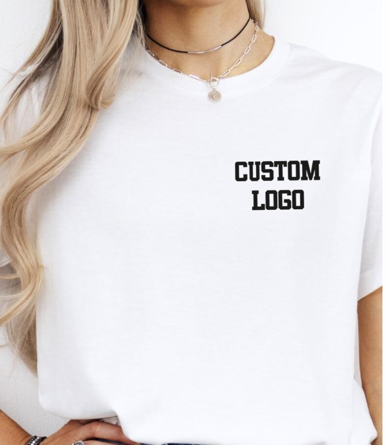 May include: White t-shirt with the text "CUSTOM LOGO" printed in black, centered on the chest. The shirt has short sleeves and a crew neck. The image shows the upper body of a person wearing the shirt.