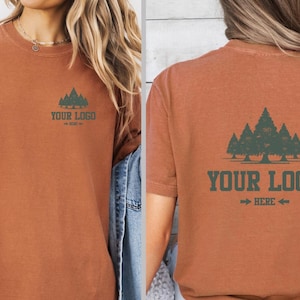 Puede incluir: Camiseta de color &oacute;xido con un gr&aacute;fico de bosque y el texto "YOUR LOGO HERE". El dise&ntilde;o se repite en la parte delantera y trasera. La camiseta tiene mangas cortas y un corte holgado.