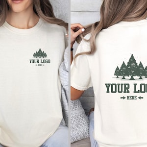 Puede incluir: Camiseta color crema con un gr&aacute;fico de bosque y el texto "YOUR LOGO HERE" en verde oscuro. El dise&ntilde;o se repite en la parte delantera y trasera. La camiseta tiene mangas cortas y un corte holgado.