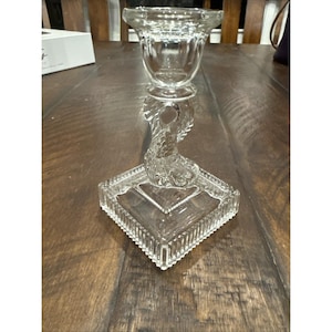 May include: A clear glass candlestick holder with a square base and a detailed, sculpted dolphin-like figure supporting the candle cup. The candlestick is designed with a decorative, textured pattern and is suitable for home decor.