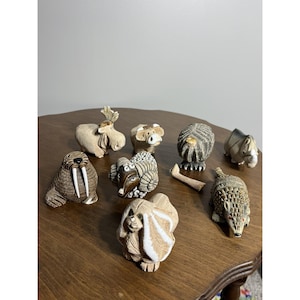 Op de afbeelding: Een verzameling kleine, gedetailleerde dierenbeeldjes, waaronder een walrus, ram en egel, gerangschikt op een houten oppervlak. De beeldjes zijn voornamelijk beige en bruin, met ingewikkelde texturen en kenmerken. De collectie omvat een verscheidenheid aan wilde dieren.