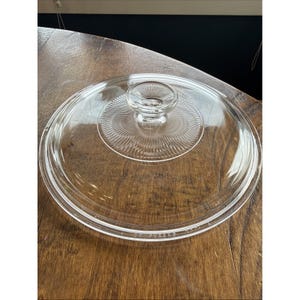 May include: A clear glass Pyrex lid with a circular design. The lid features a central knob and a series of concentric ridges radiating outwards. The rim of the lid is slightly curved. The lid is sitting on a wooden surface.