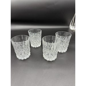 May include: Four clear, patterned drinking glasses. The glasses have a diamond-like design and are arranged on a dark surface. The glasses are empty and appear to be made of glass.
