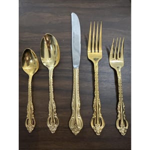 Puede incluir: Un juego de cubiertos dorados, que incluye una cuchara, una cuchara sopera, un cuchillo y dos tenedores. Los utensilios tienen mangos ornamentados y detallados y están dispuestos sobre una superficie de madera oscura. El cuchillo tiene una hoja plateada.