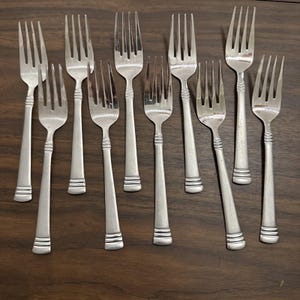 May include: A collection of ten silver forks arranged on a dark wooden surface. Each fork has a long handle with a decorative band near the base and a four-tine head. The forks are evenly spaced and angled, creating a visually appealing arrangement.
