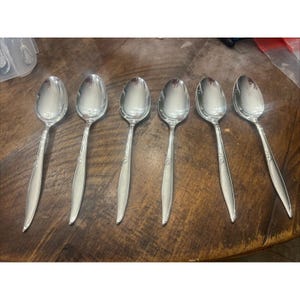 May include: Six silver spoons displayed on a wooden surface. The spoons have a traditional design with a decorative pattern on the handle. The spoons are reflective, showing the surrounding environment.