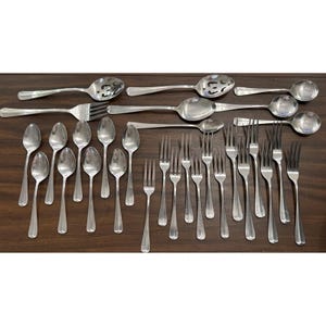 May include: A collection of stainless steel silverware, including spoons, forks, and serving spoons, arranged on a dark wooden surface. The spoons vary in size, with some having slotted designs. The forks have four tines.