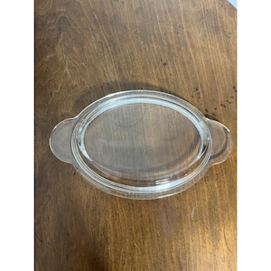 Può includere: Piatto ovale in vetro trasparente con due manici laterali. Il piatto è trasparente, consentendo una visione della superficie sottostante. Il piatto è probabilmente per servire o cuocere.