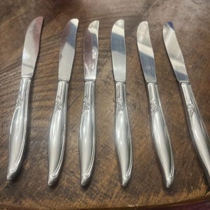 May include: A collection of six silver butter knives. Each knife features a curved blade and a decorative handle with a floral pattern. The knives are displayed on a wooden surface.