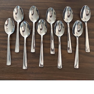 May include: A set of nine polished stainless steel spoons arranged in two rows. Each spoon features a smooth, reflective oval bowl and a tapered handle. The metal surface of each spoon shows a clear reflection of the surrounding environment.