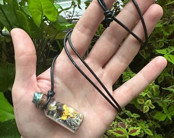 Könnte beinhalten: Eine kleine Glasflasche gefüllt mit getrockneten Blumen und Kräutern hängt an einer schwarzen Lederschnur. Die Flasche hat einen kupferfarbenen Deckel und ist gefüllt mit getrockneten gelben, weißen und braunen Blumen.