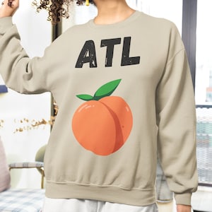 May include: Beige sweatshirt with the text "ATL" in black block letters above a cartoon peach with green leaves. The sweatshirt is a crew neck style.