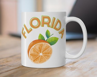 Puede incluir: Taza de cerámica blanca con un gráfico de una rodaja de naranja y la palabra "FLORIDA" en letras naranja y verde.