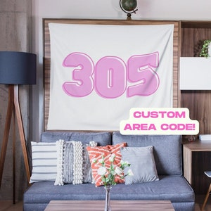 May include: A white tapestry featuring a pink 305 area code design. The tapestry is displayed on a wall in a living room setting, with a sofa, cushions, and a lamp. The text "CUSTOM AREA CODE!" is written on a white sticker below the design.