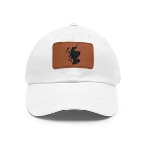 May include: A white baseball cap featuring a rectangular brown leather patch centered on the front panel. The patch displays a black silhouette of the map of Scotland, showcasing the country's distinct coastal outline and islands in a dark contrast.