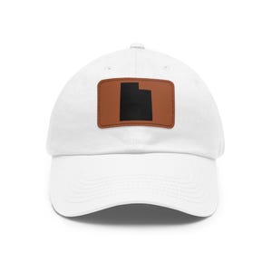 Pode incluir: Um boné de beisebol branco com um emblema retangular de couro castanho centrado na parte frontal. O emblema apresenta uma silhueta preta sólida do estado do Utah. O boné tem uma aba curva e uma copa estruturada com ilhós costurados.