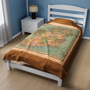 May include: A brown fleece blanket with an antique map of Ireland printed on it. The map is in shades of green, yellow, and blue. The blanket is folded and displayed on a white bed frame.