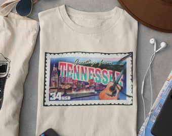 May include: A beige t-shirt with a vintage-style postcard graphic featuring a cityscape and the text "Greetings from Tennessee". The postcard has a red, blue, and green color scheme and a guitar in the bottom right corner.