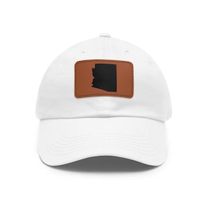May include: A white baseball cap featuring a rectangular brown patch centered on the front. The patch displays a solid black silhouette of the state of Arizona. The cap has a curved brim and a structured crown with a button at the top center.