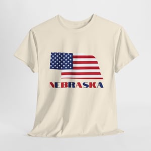 Op de afbeelding: Een crèmekleurig T-shirt met een afbeelding van de staat Nebraska in de vorm van de Amerikaanse vlag. De tekst "NEBRASKA" is in blauwe en rode letters onder de vlag gedrukt.
