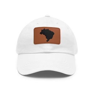 Puede incluir: Una gorra de béisbol blanca con un parche rectangular de cuero marrón centrado en el panel frontal. El parche muestra una silueta negra del mapa de Brasil, cosida sobre la tela con hilo visible a lo largo de los bordes del parche.