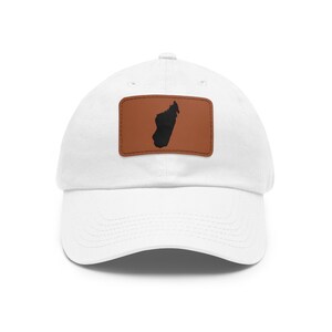 May include: A white baseball cap featuring a rectangular brown patch on the front. The patch displays a black silhouette of the island of Madagascar centered within its borders. The cap has a curved brim and a structured crown design.