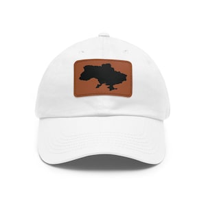 May include: A white baseball cap featuring a rectangular brown leather patch centered on the front. The patch displays a solid black silhouette of the map of Ukraine. The cap has a curved brim and a structured crown with stitched eyelets.