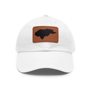 May include: A white baseball cap featuring a rectangular brown patch on the front. The patch displays a solid black silhouette map of Honduras centered on the material. The cap has a curved brim and a structured crown with a button on top.