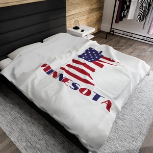 Puede incluir: Una manta blanca con un diseño de bandera estadounidense roja, blanca y azul en forma del estado de Minnesota. Las palabras "MINNESOTA" están impresas en azul en la manta.