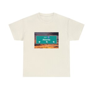 May include: A white t-shirt featuring a green road sign that reads "East I-20 Atlanta" with arrows pointing downwards. The sign is set against a sunset backdrop.