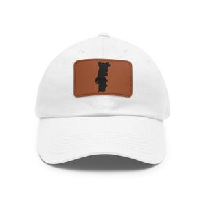 May include: A white baseball cap featuring a rectangular brown leather patch centered on the front panel. The patch displays a black silhouette of the country of Portugal. The cap has a curved brim and a structured crown with stitched eyelets.