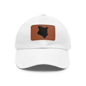 May include: A white baseball cap featuring a rectangular brown leather patch centered on the front. The patch displays a solid black silhouette of the map of Kenya. The cap has a curved brim and a structured crown with stitched eyelets.