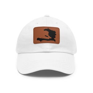 Puede incluir: Una gorra de béisbol blanca con un parche rectangular marrón centrado en la parte delantera. El parche muestra una silueta negra de la isla de La Española, específicamente el mapa de Haití, con costuras visibles a lo largo de los bordes del parche.