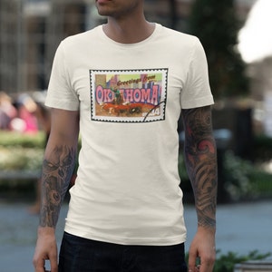 Puede incluir: Camiseta blanca con un gráfico de postal vintage que muestra a un vaquero montando un toro con el texto "Greetings from OKLAHOMA" y un sello rojo, blanco y azul.