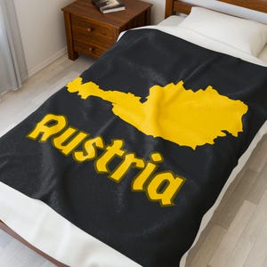 May include: A black blanket with a yellow outline of Austria and the word "Austria" in yellow. The blanket is on a bed with a white pillow and a wooden nightstand in the background.