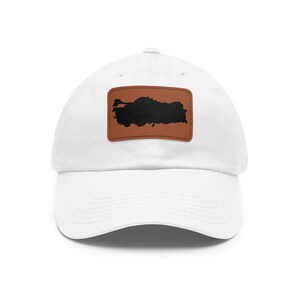May include: A white baseball cap featuring a rectangular brown patch centered on the front. The patch displays a solid black silhouette of the map of Turkey. The cap has a curved brim and a structured crown with a button at the top center.