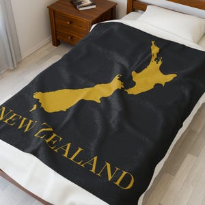 May include: A black blanket featuring a gold outline of New Zealand and the words "NEW ZEALAND" in gold. The blanket is on a bed with a white pillow and a wooden bedside table.