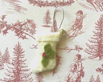 May include: A small, off-white felt Christmas stocking ornament with a green holly leaf and red berry detail. The ornament has a silver loop for hanging and a small gold bell. The background features a red and white winter scene with the text "MERRY CHRISTMAS".