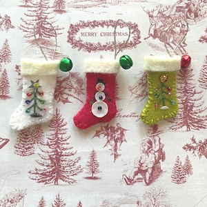 May include: Three small felt Christmas stockings with hanging loops and jingle bells. One white stocking features a beaded tree, one red stocking has a button snowman, and one lime green stocking displays a beaded tree design with embroidery.