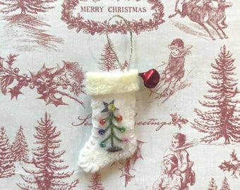 May include: A white felt Christmas stocking ornament featuring a hand-stitched green tree design decorated with small colorful beads and a silver star at the top. A silver hanging loop is attached to the top, and a small red bell hangs from the side.