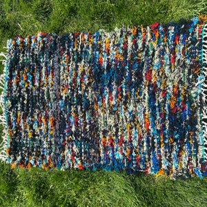 Puede incluir: Una alfombra colorida tejida a mano con una superficie texturizada y peluda. La alfombra presenta una mezcla de colores azules, rojos, amarillos, blancos y negros en un patrón a rayas.