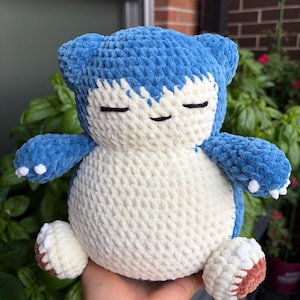May include: A plush crochet toy shaped like a Snorlax character, featuring a soft blue and cream-coloured yarn texture. The figure has a round body, small ears, closed eyes, a tiny mouth, and white claws on its hands and feet with brown soles.