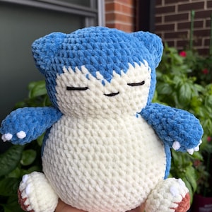 May include: A plush toy crafted from blue and cream-colored yarn, featuring a rounded body, small ears, and closed eyes. The creature has a cream-colored belly and face, with blue limbs and small white claws on the hands and feet, and brown soles.