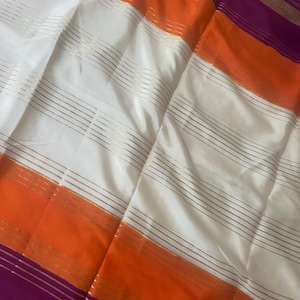 May include: A close-up of a silk fabric with horizontal stripes. The fabric features alternating bands of white, orange, and purple. Thin gold lines run across the white sections. The fabric has a shiny, smooth texture.