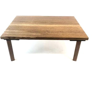 May include: A small, square wooden table with a natural wood grain finish. The table has four dark brown legs. The tabletop is approximately 24 inches square and 12 inches high.